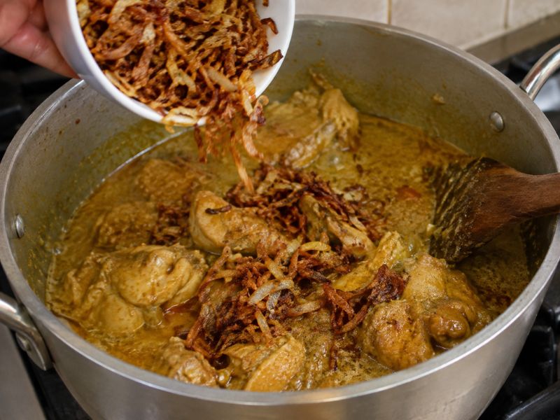 Golden fried onions being added to creamy chicken korma in a cooking pan.