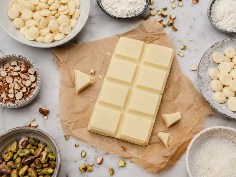 Homemade white chocolate bar surrounded by cocoa butter, milk powder, chopped almonds, pistachios, coconut, and white chocolate pieces on parchment paper.