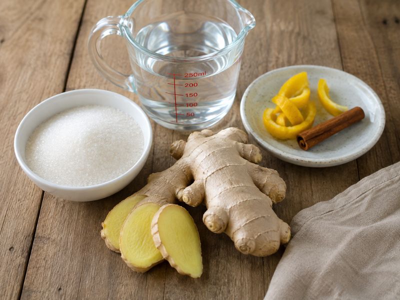 Fresh ginger root and sliced  arranged with white sugar, water in a measuring jug, lemon peel, and a cinnamon stick on a rustic wooden table.