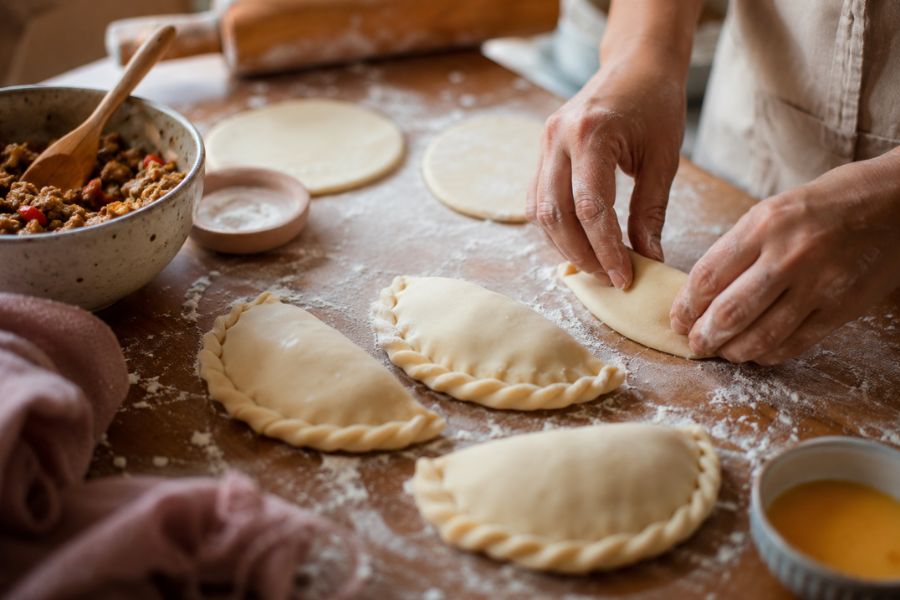 Hands sealing homemade empanada dough on a floured wooden countertop with rolling pin, egg wash, and filling for perfect flaky empanadas.