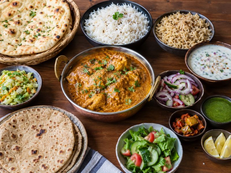 Chicken korma served with naan, roti, basmati rice, jeera rice, raita, salad, chutney, pickles, and lemon wedges on a wooden table.